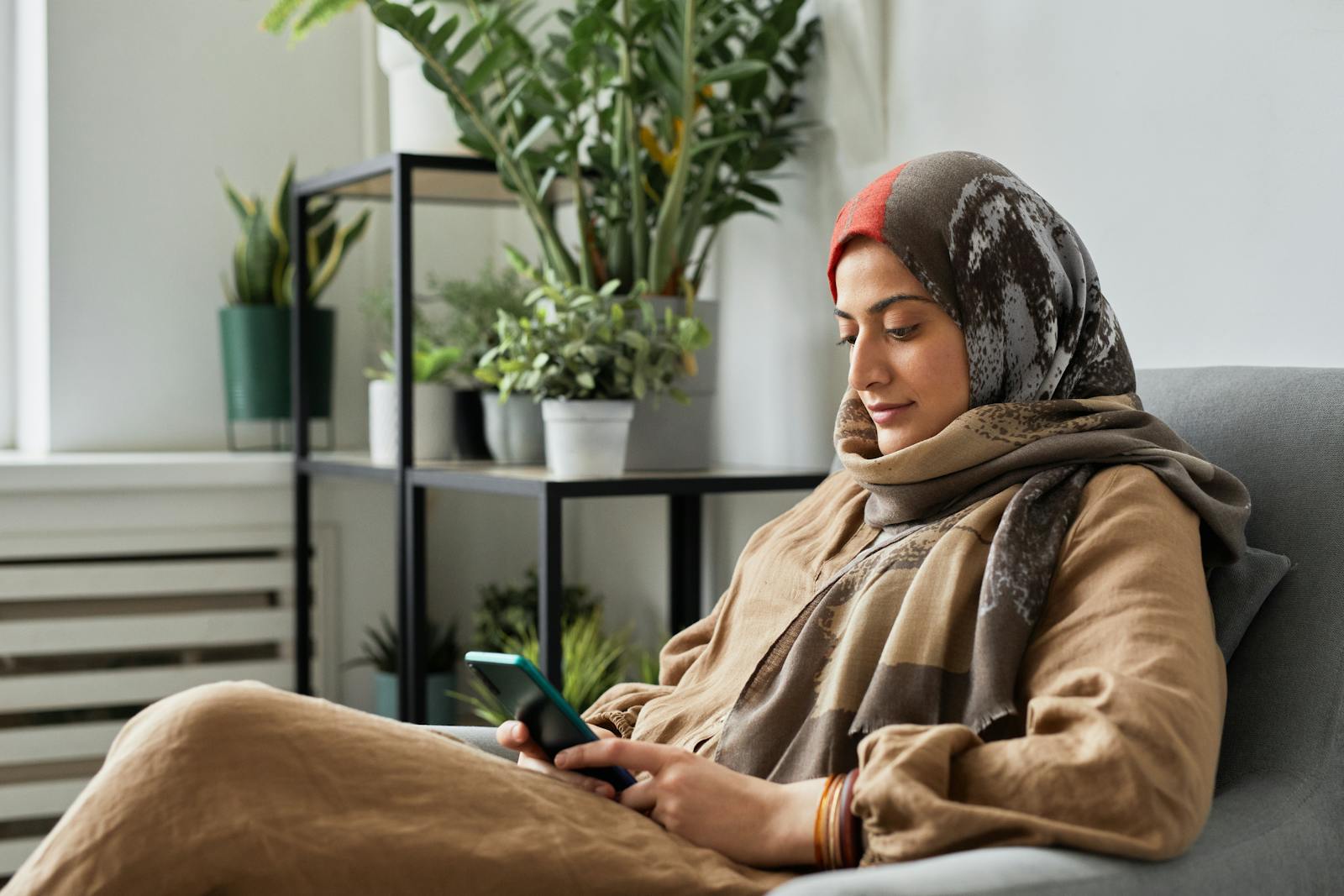 A Muslim woman wearing a headscarf enjoys phone time while relaxing indoors.